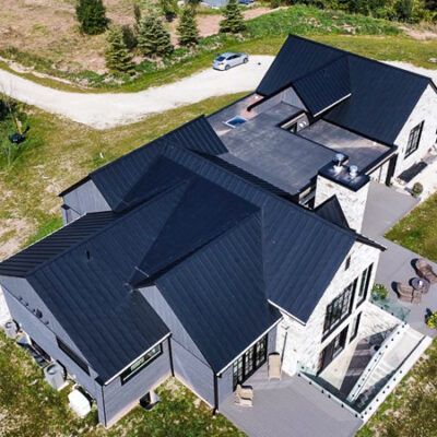 Aerial view of a modern house with a dark metal roof, gray siding, white stone accents, and a patio, set in a grassy area.
