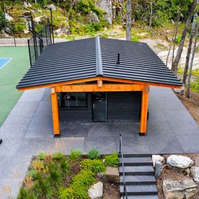 A modern wood-framed pavilion with a dark roof sits on a stone patio next to a tennis court and outdoor stairs.