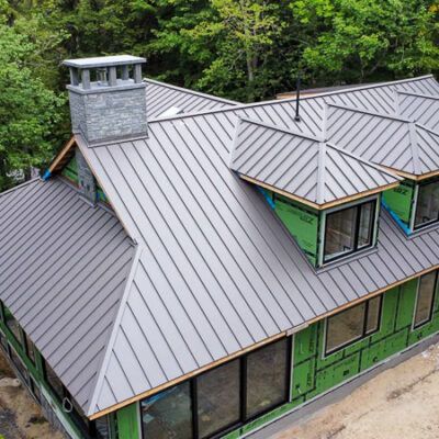 An aerial view of a house under construction with a grey standing-seam metal roof, chimney, and green sheathing.