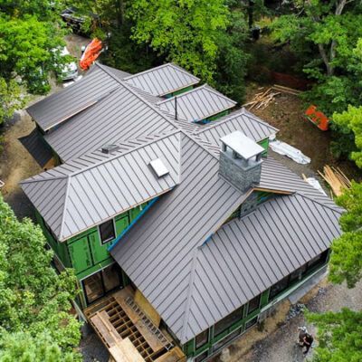 An aerial view of a house under construction with a gray metal roof, green sheathing, and a stone chimney.