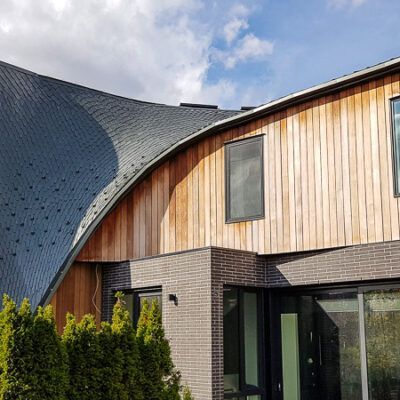 Modern house exterior with a curved, shingled roof, light wood siding, brick ground floor, and large glass windows.