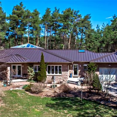 A single-story, stone-walled house with a dark metal roof, set against a backdrop of tall pine trees on a sunny day.