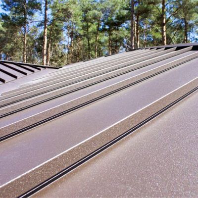 A close-up of a brown, standing seam metal roof set against a backdrop of pine trees.