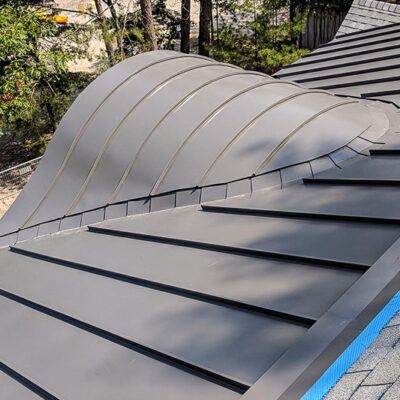 A close-up view of a matte grey, curved metal roof section transitioning into a flat, standing seam metal roof.