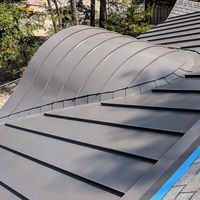 A close-up view of a matte grey, curved metal roof section transitioning into a flat, standing seam metal roof.