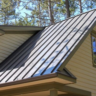 A dark metal standing-seam roof on a house with beige siding and a porch, surrounded by pine trees under a clear sky.