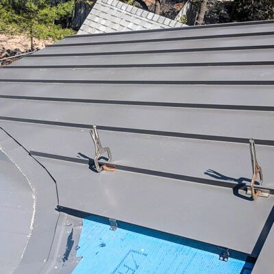 Metal roofing panels are being installed on a roof, secured with two clamps over blue underlayment.