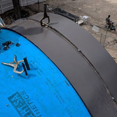 A metal clamp secures two gray roofing panels together over a blue underlayment on an unfinished construction project.