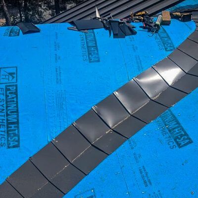 A residential roof under construction, featuring blue synthetic underlayment and a diagonal row of installed roof tiles.