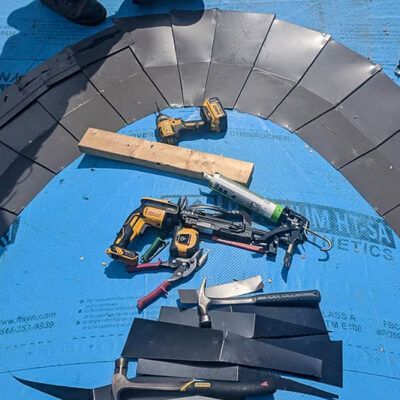 A top-down view of roofing tools, including a nail gun, drill, and hammer, resting on blue underlayment near curved tiles.