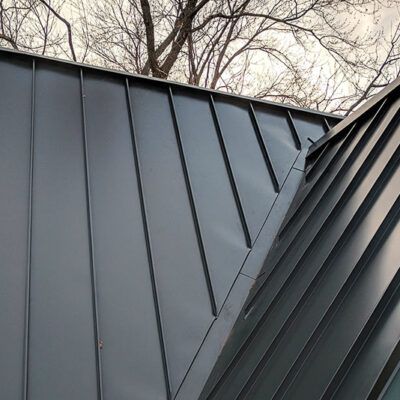 A close-up of a modern dark gray metal standing seam roof with a central valley meeting tree branches against the sky.