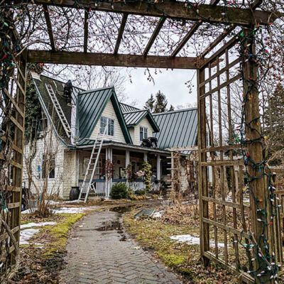 Roofers work on a metal-roofed house viewed through a wooden arbor, with a stone path leading toward the front porch.