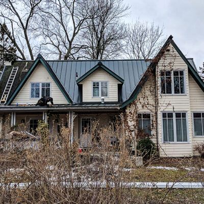 A house with cream siding, a gray metal roof, and gabled windows, under maintenance with a ladder leaned against it.