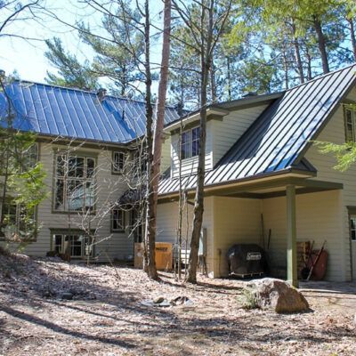 A two-story, light yellow house with a dark blue metal roof nestled among trees in a wooded yard with scattered leaves.