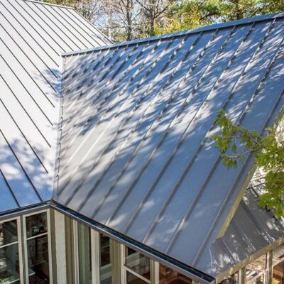A high-angle view of a modern house roof with grey standing-seam metal panels, showing shadows cast by nearby trees.