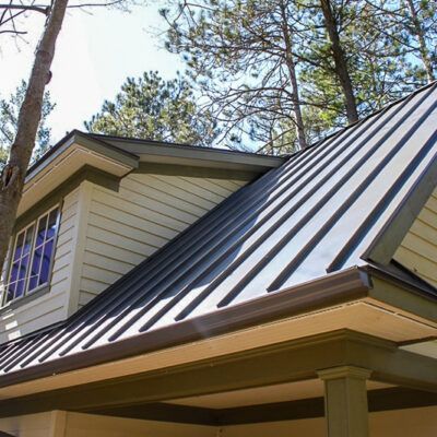 A home with a dark gray standing seam metal roof, beige siding, and a covered porch, set against a backdrop of pine trees.