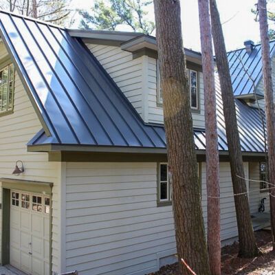 Light-colored house with a dark metal roof, garage door, and windows, set among trees.