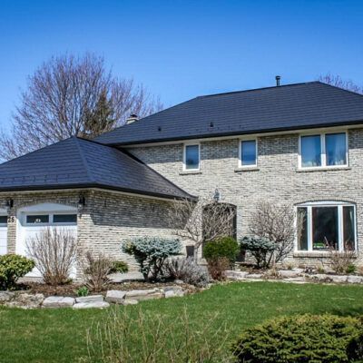 Two-story stone house with a dark gabled roof, white garage doors, and landscaped lawn under a clear blue sky.