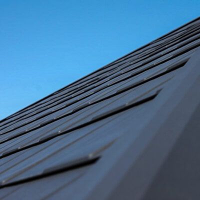 A low-angle view of a dark metal shingle roof against a clear, bright blue sky.