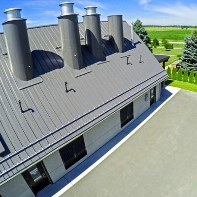 Aerial view of a gray metal building with four cylindrical chimneys on its slanted roof, near a grassy field.