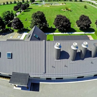 An aerial view of a gray metal roof on a building, featuring four chimneys, with a green lawn and trees in the background.