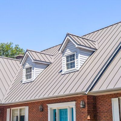 A red brick house with a light gray metal roof featuring two dormer windows and a blue front door.