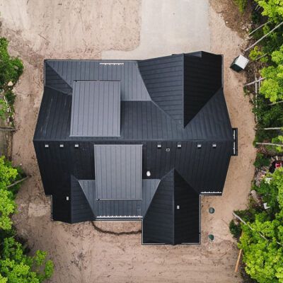 Aerial view of a house with a dark metal roof, surrounded by trees and a dirt yard.
