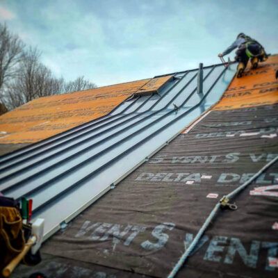 A worker installs gray standing seam metal roofing on a residential slope over black underlayment.