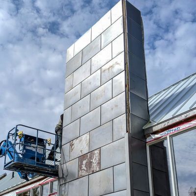 A construction worker in a blue aerial lift works on a metal-clad chimney tower attached to a building.
