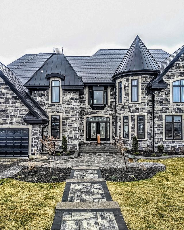 A large, two-story stone house with a dark metal roof, a prominent turret, and a paved walkway leading to the front door.