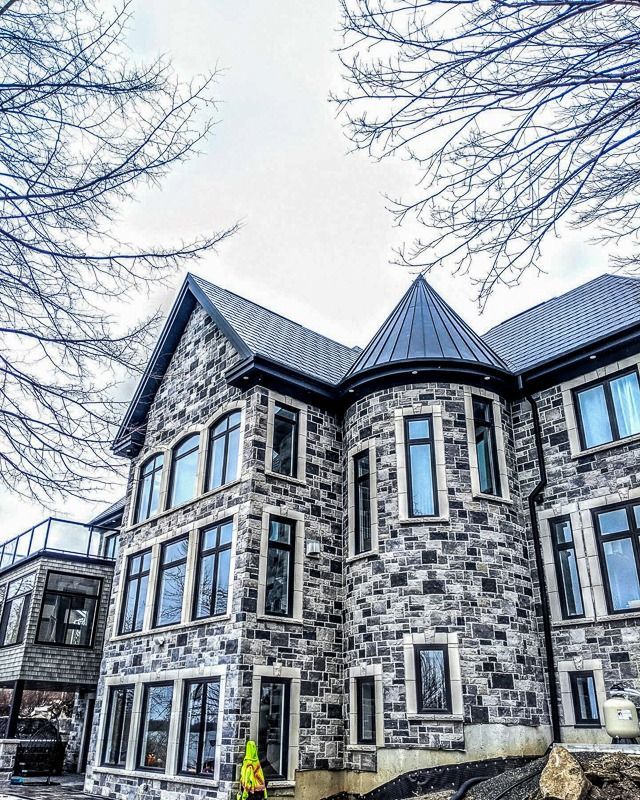 A modern, multi-story stone house with a turret and dark shingled roof, framed by bare tree branches against a bright sky.