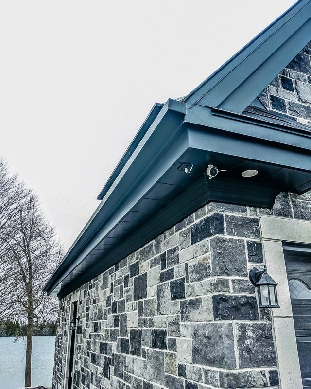 A security camera mounted under the dark gray metal roof overhang of a house with a stone exterior wall.