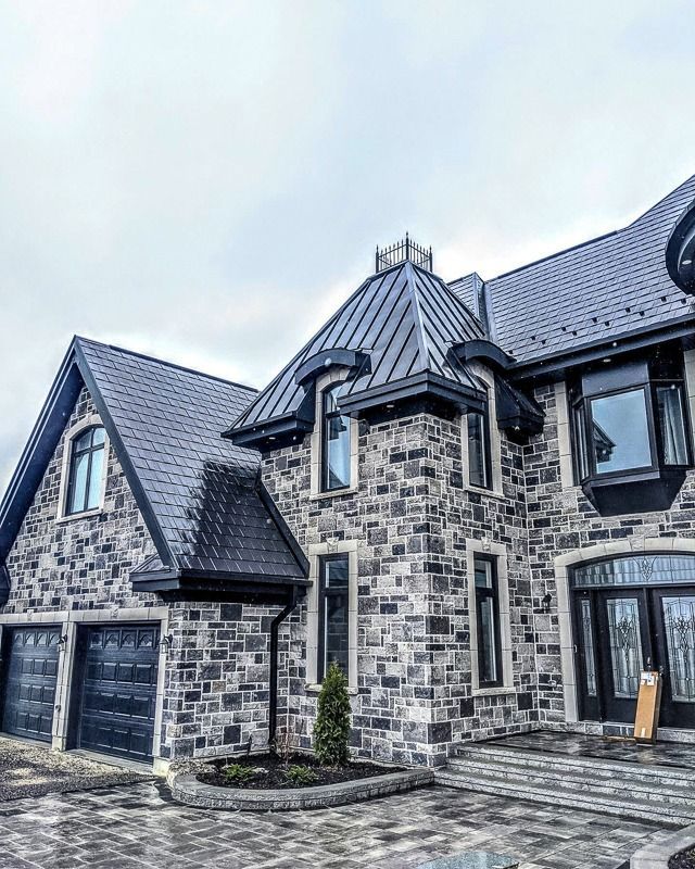 A large two-story stone house with a complex shingled roof, dark trim, multiple windows, and a paved driveway.