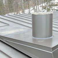 A gray metal roof with standing seams features a circular chimney vent sealed with light-colored flashing.