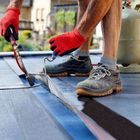 A worker in red gloves uses a propane torch to seal roofing material on a flat roof.