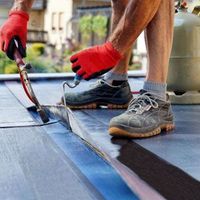A worker in red gloves uses a propane torch to seal roofing material on a flat roof.