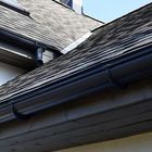 A low-angle view of a grey shingled roof with dark gutters and a metal flashing valley against a clear blue sky.