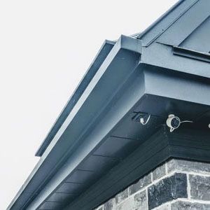 Low-angle view of a dark metal roof corner and soffit with attached security camera and wall-mounted hook.