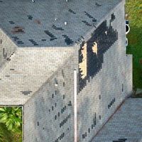 Aerial view of a damaged roof with missing shingles, exposing the underlying wooden deck in a large patch.