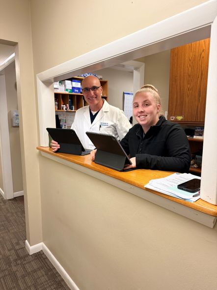 Two staff members at a reception counter with open laptops in a hallway pass-through window.