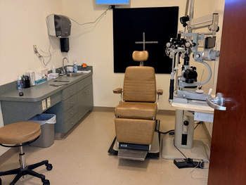 Eye exam room with exam chair, slit lamp, desk, stool, and wall-mounted monitor