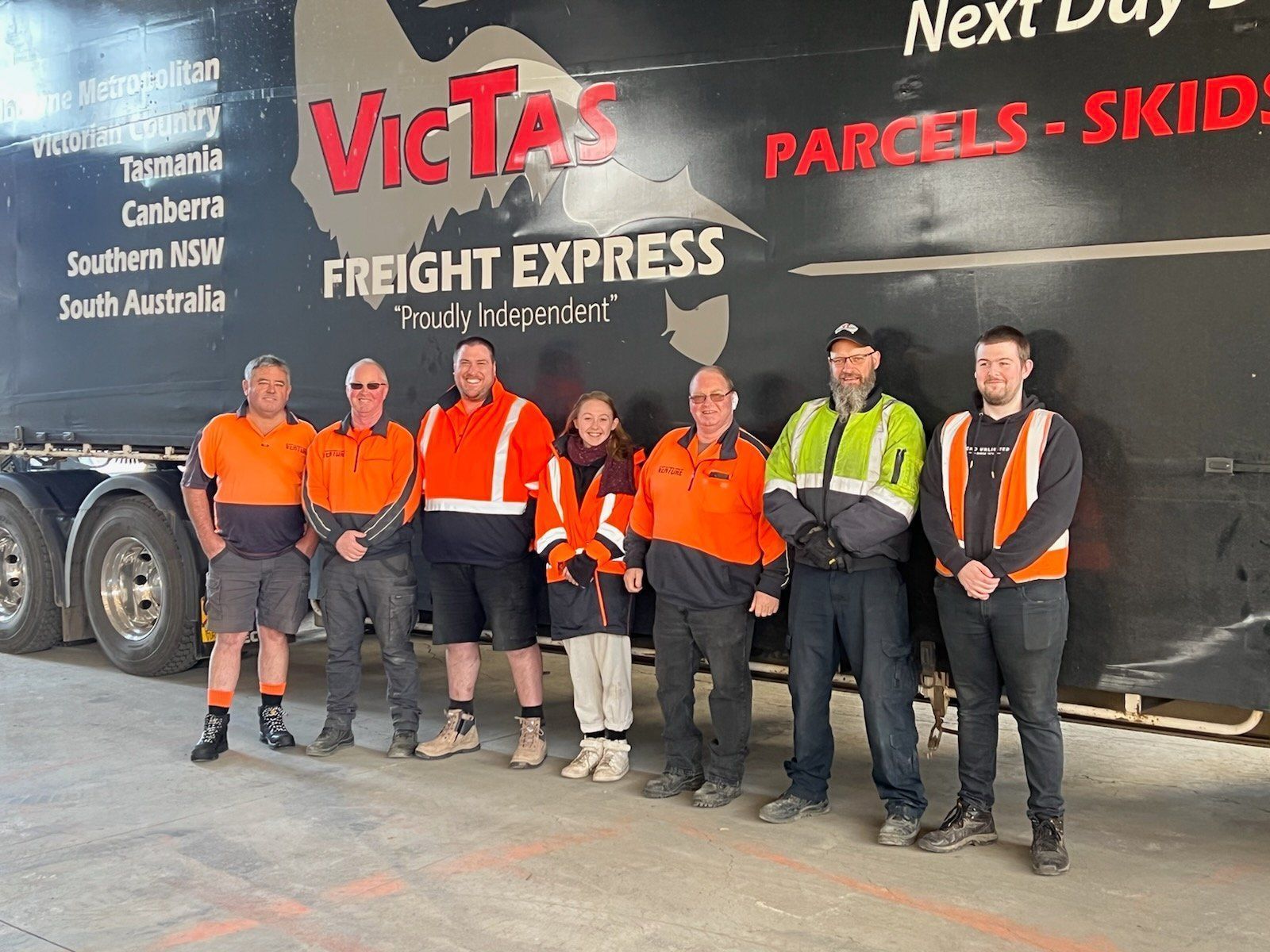 Six workers in hi-vis gear standing in front of a VicTas Freight Express truck mural.