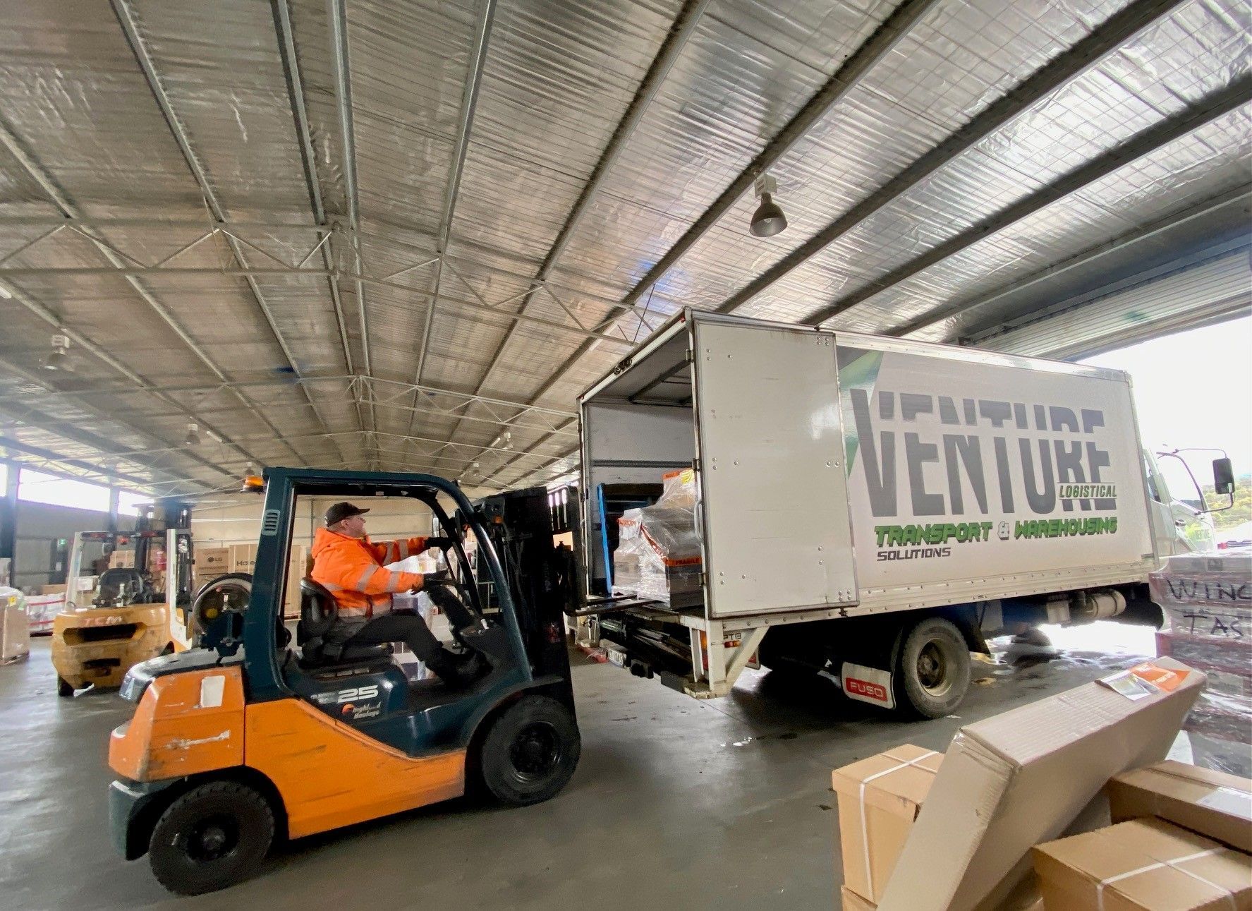Forklift loading a white delivery truck in a warehouse, with an orange-vested worker at the wheel.