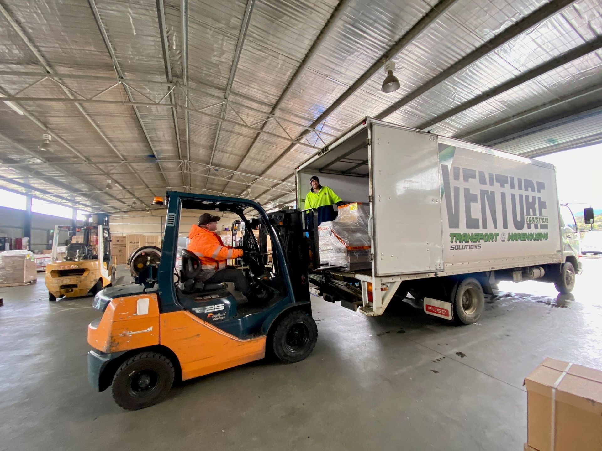Orange forklift unloading a white truck in a warehouse, with a worker nearby Orange forklift unloading a white truck in a warehouse, with a worker nearby