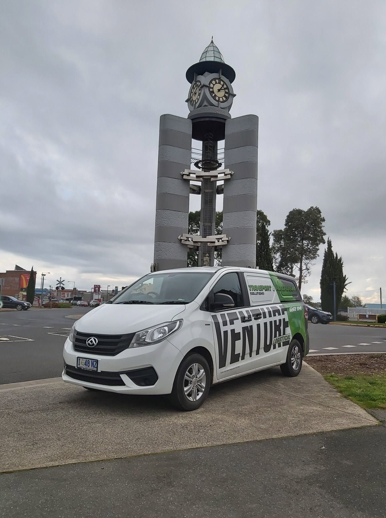 White van parked by a large clock monument under a cloudy sky.