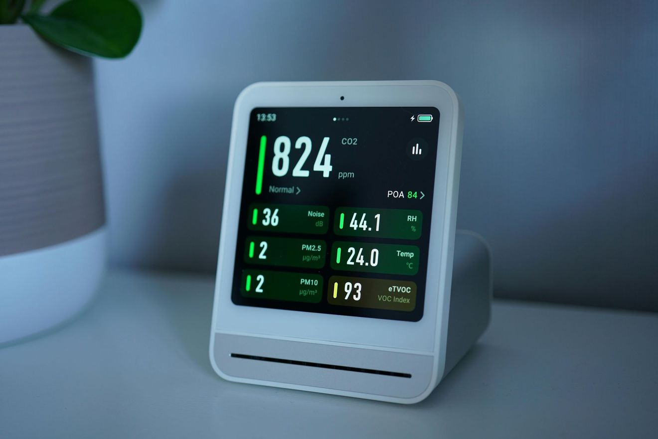 A white air quality monitor displays various readings in green on a small screen, sitting on a white surface.
