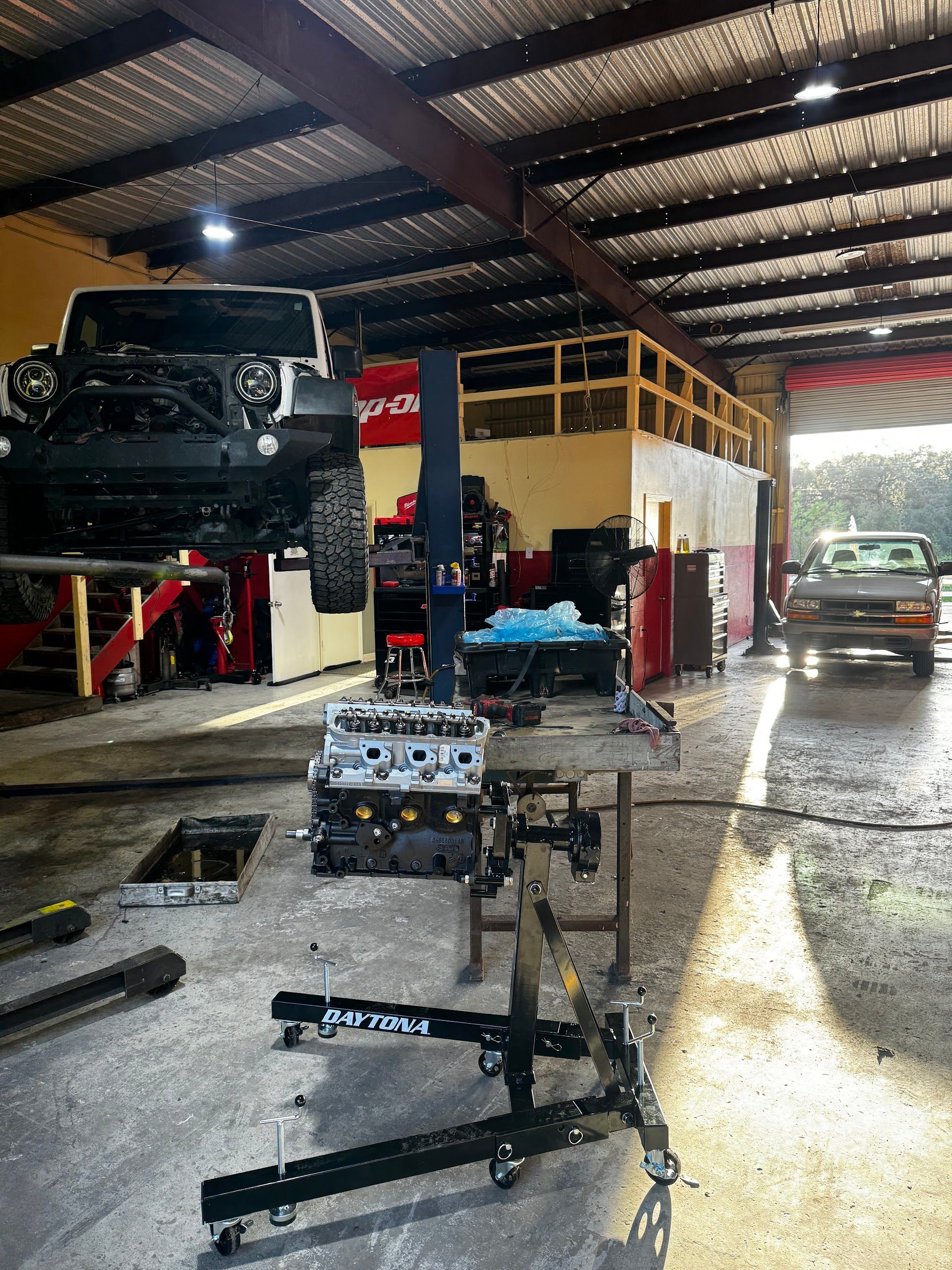 An auto repair shop with a Jeep lifted on a rack, an engine on a hoist, and a truck parked near the open bay door. | Keep It Rollin’ Auto Repair LLC