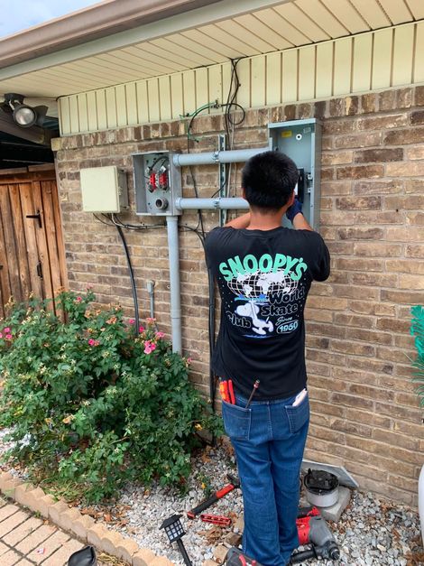 Person standing by an outdoor electrical panel on a brick wall, working with tools in a yard.