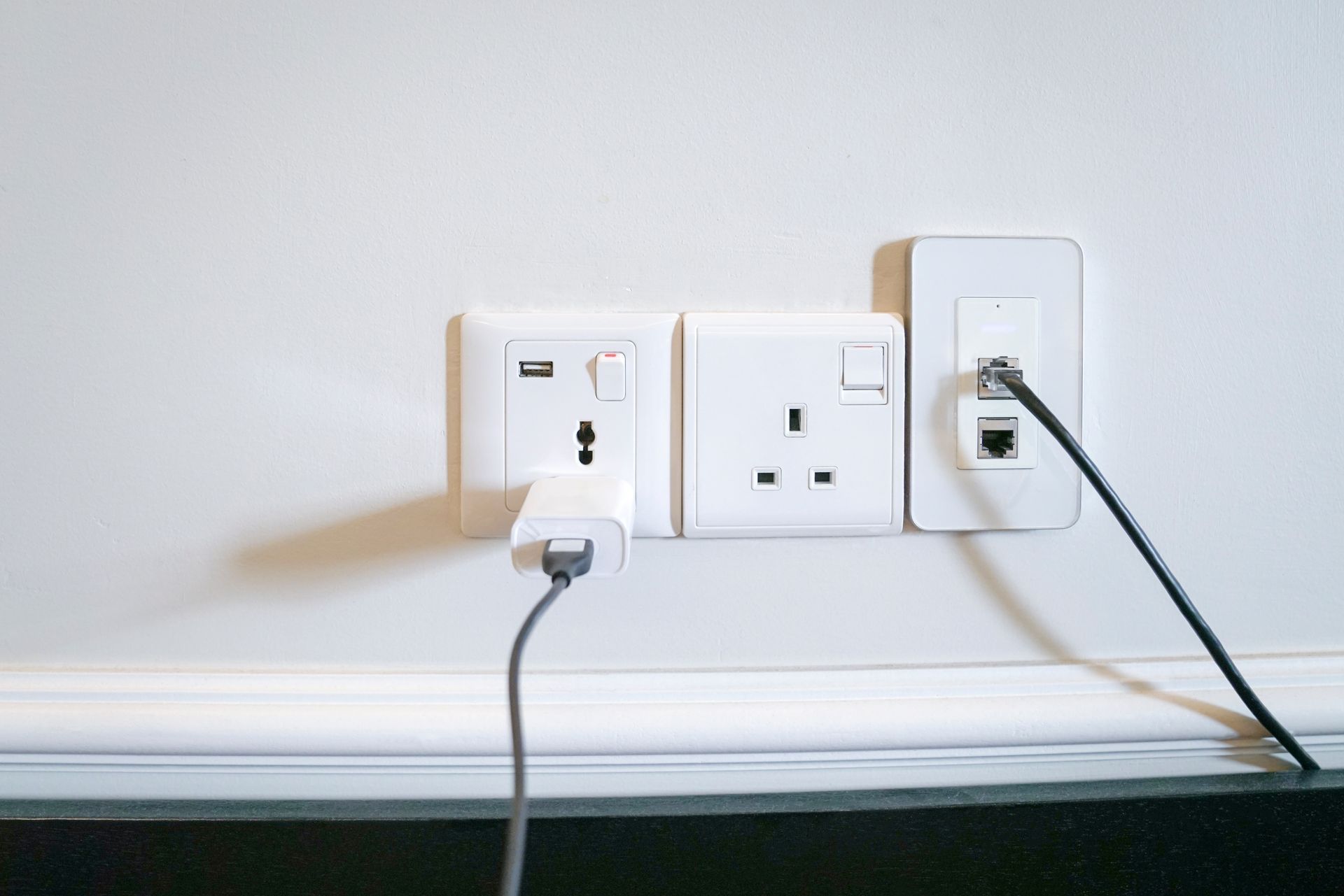 Two wall outlets with white and black plugs on a white wall above a baseboard