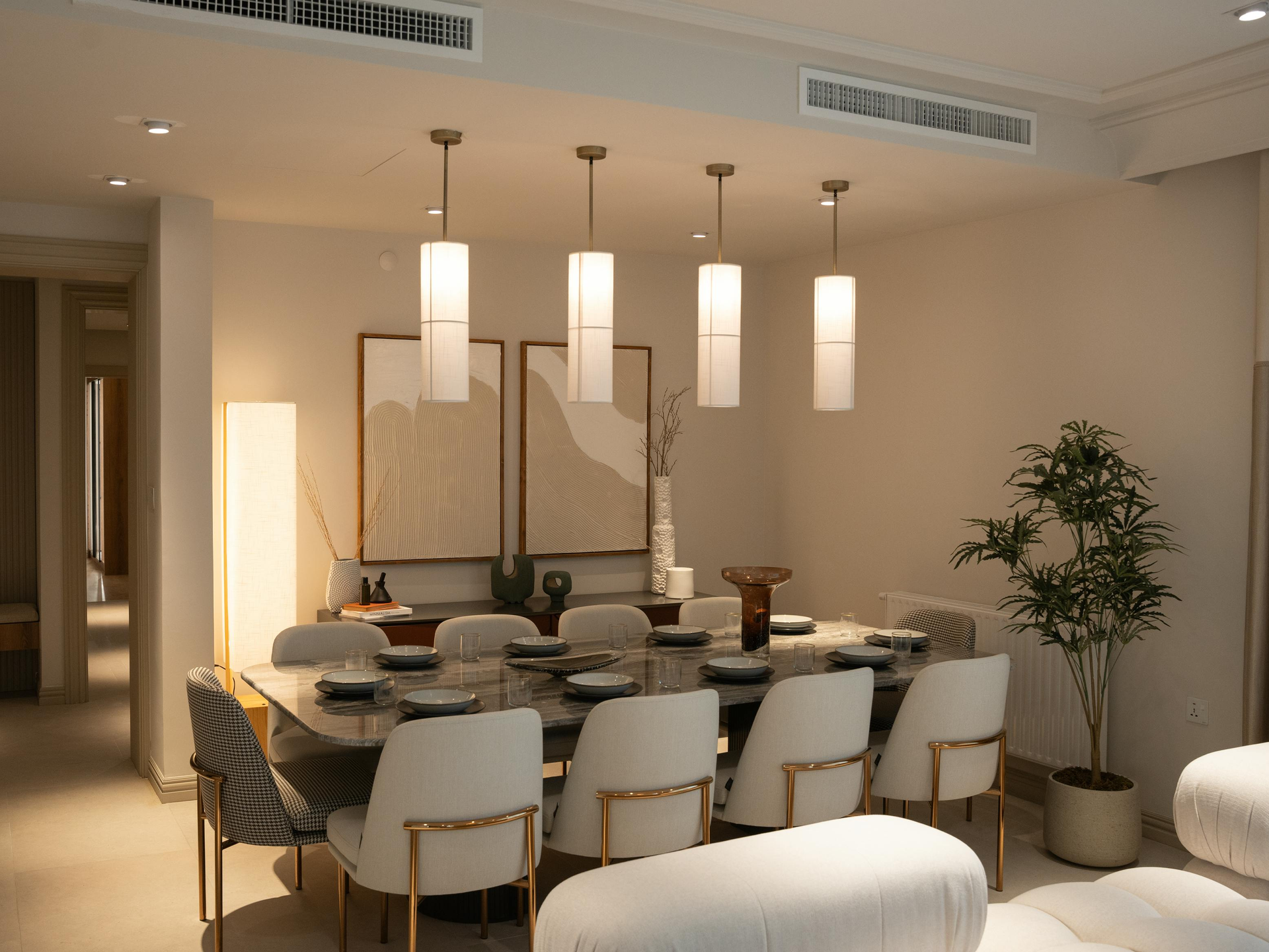 Modern dining room with a wooden table, white chairs, pendant lights, and a potted tree by the wall